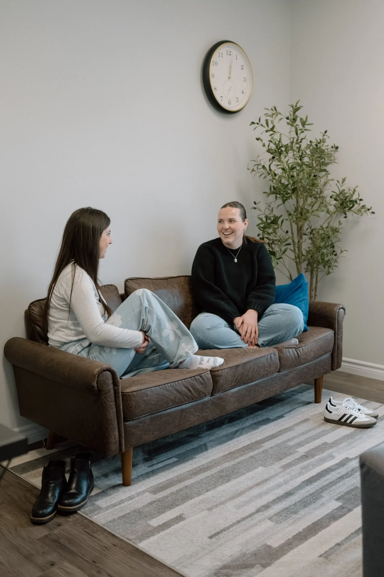Two women sitting on a brown sofa in a living room, smiling and talking. One is wearing pajamas with slippers, and the other is dressed casually. A pair of sneakers is on the floor, a wall clock is on the wall, and a green potted plant is beside them.