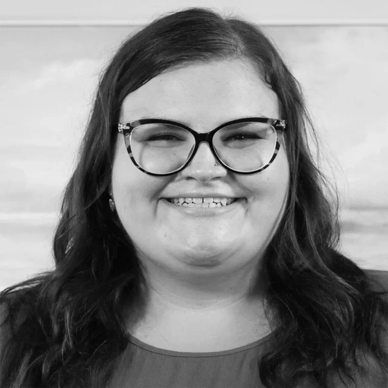 A woman with dark, wavy hair wearing glasses smiling at the camera in a black and white photo.