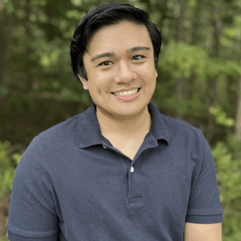 A young man with black hair, smiling, wearing a navy blue polo shirt, outdoors with green trees in the background.