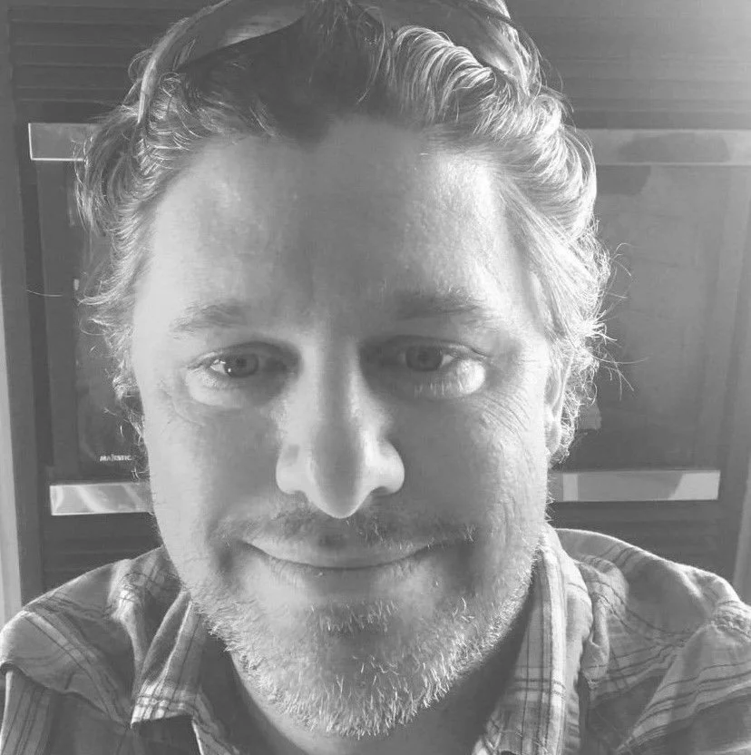 A close-up black and white portrait of a man with wavy hair, light facial hair, and a gentle smile.