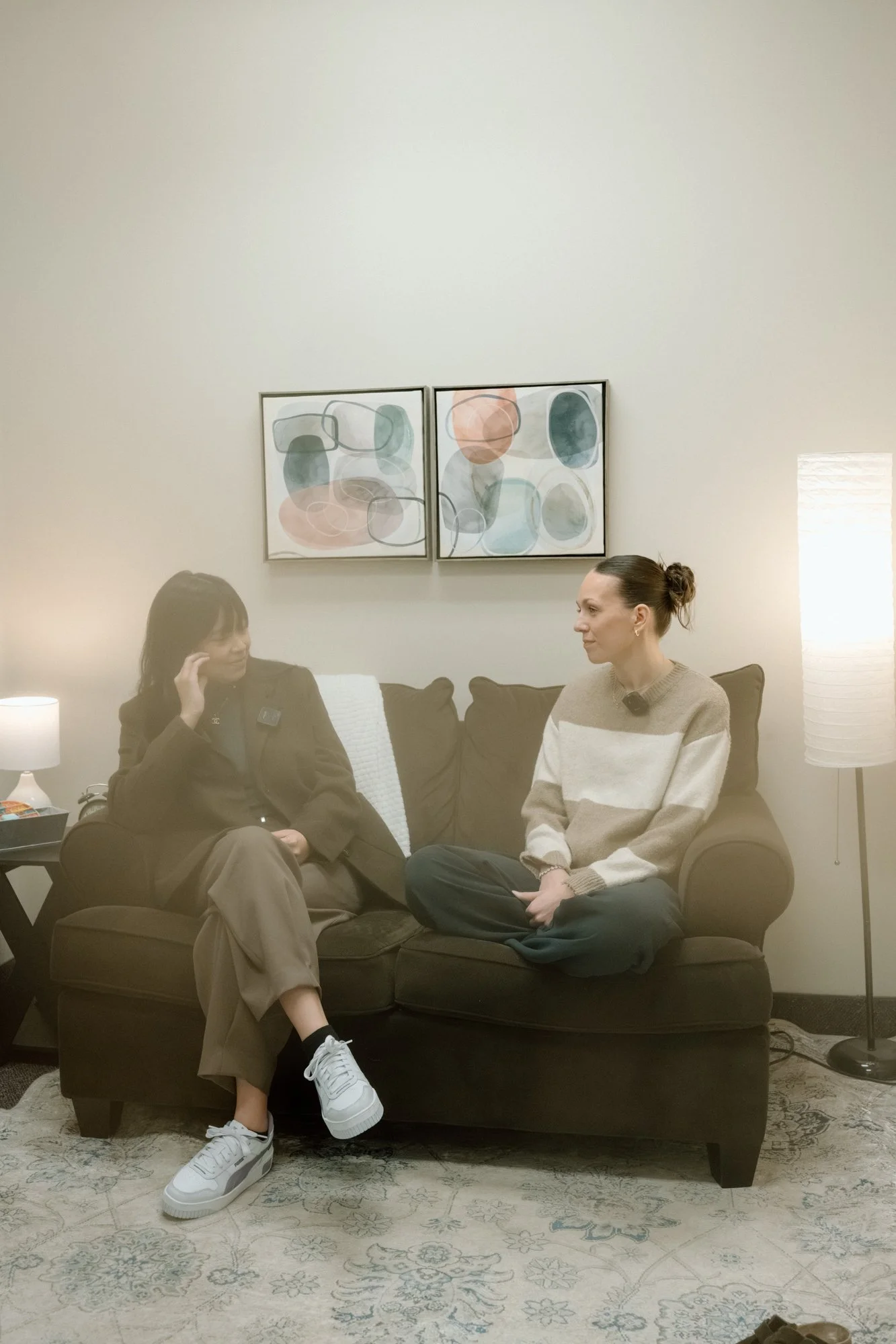 Two women sitting on a black sofa in a living room, engaged in conversation. The woman on the left is wearing a dark jacket and beige pants, with white sneakers. The woman on the right is wearing a beige and white striped sweater and dark pants, sitting cross-legged. Behind them, there is wall art with abstract shapes and colors. A small side table with a lamp and other items is to the left, and a tall lamp with a white shade is on the right. The floor has a patterned rug.
