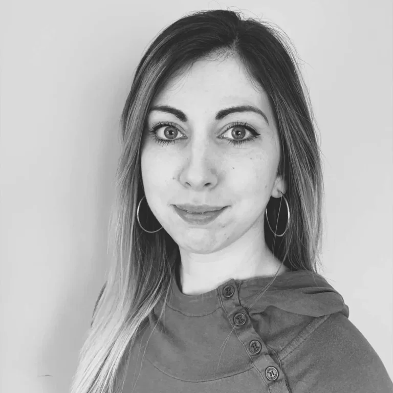 A black and white portrait of a young woman with long hair, wearing hoop earrings and a top with buttons on the shoulder, looking at the camera with a slight smile.