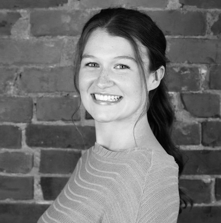 Black and white photo of a smiling woman with long hair standing in front of a brick wall, wearing a striped top.