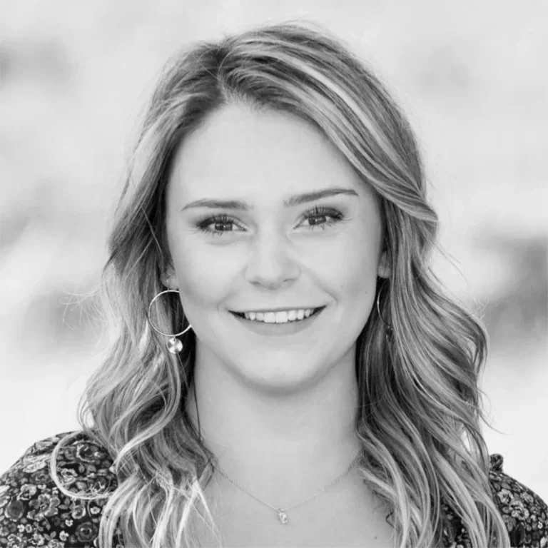 A young woman with long wavy hair smiling, wearing hoop earrings and a floral top, in black and white photograph.