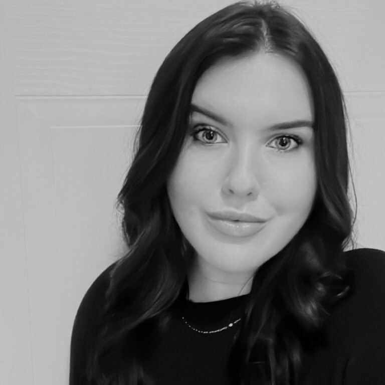 Black and white photo of a young woman with long wavy hair, smiling gently, wearing a black top and a delicate necklace.