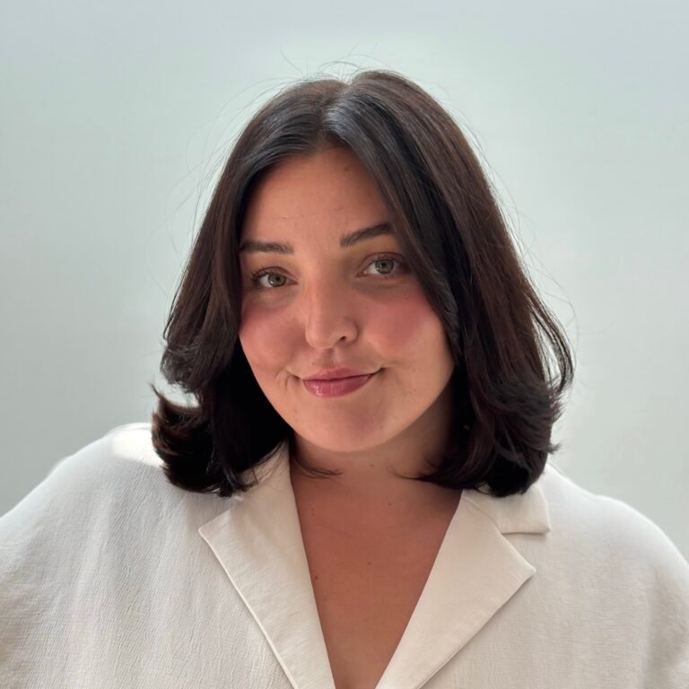 A woman with shoulder-length dark hair and light skin wearing a white blazer, smiling slightly towards the camera against a plain light green background.