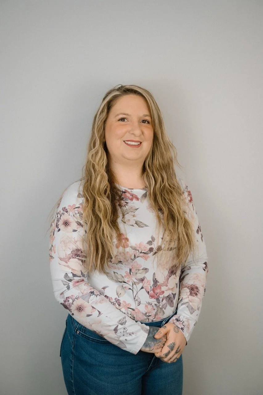 A woman with long, wavy blonde hair, wearing a white floral long-sleeve shirt and blue jeans, smiling and standing against a plain light gray wall.