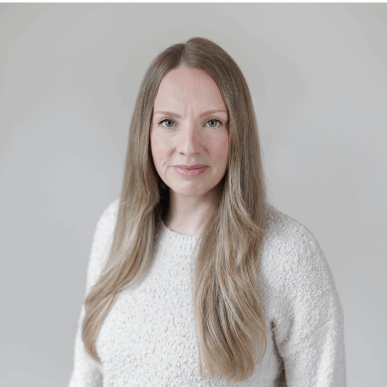 A woman with long, light brown hair wearing a white textured sweater, looking at the camera with a neutral expression against a plain light gray background.