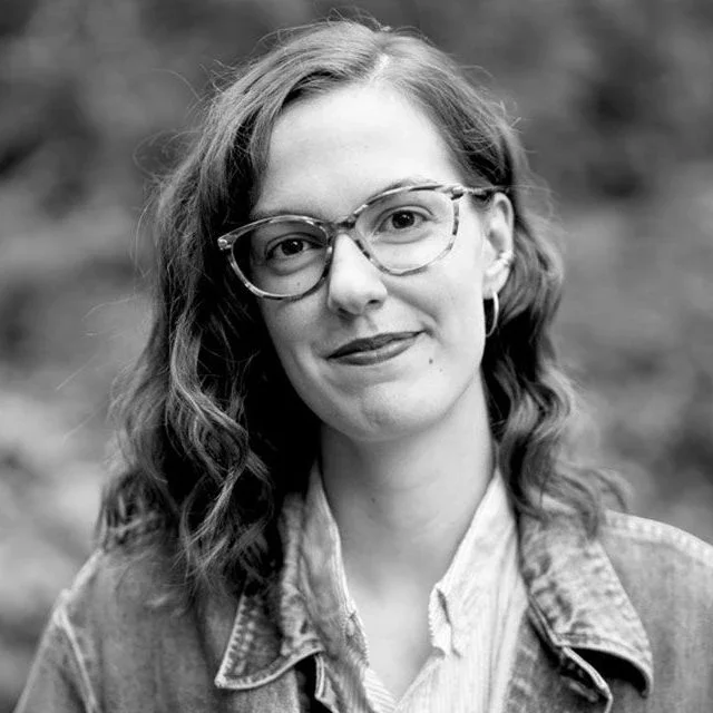 Black and white portrait of a young woman with wavy hair, wearing glasses, earrings, a collared shirt, and a denim jacket, standing outdoors.