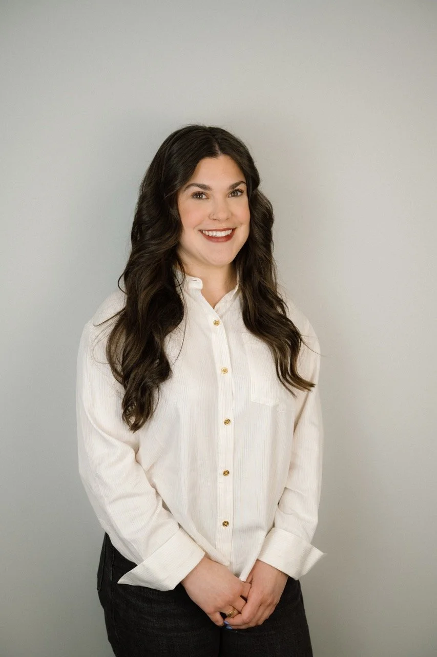 A woman with long dark wavy hair, wearing a white button-up shirt with gold buttons, smiling and standing against a light gray background.