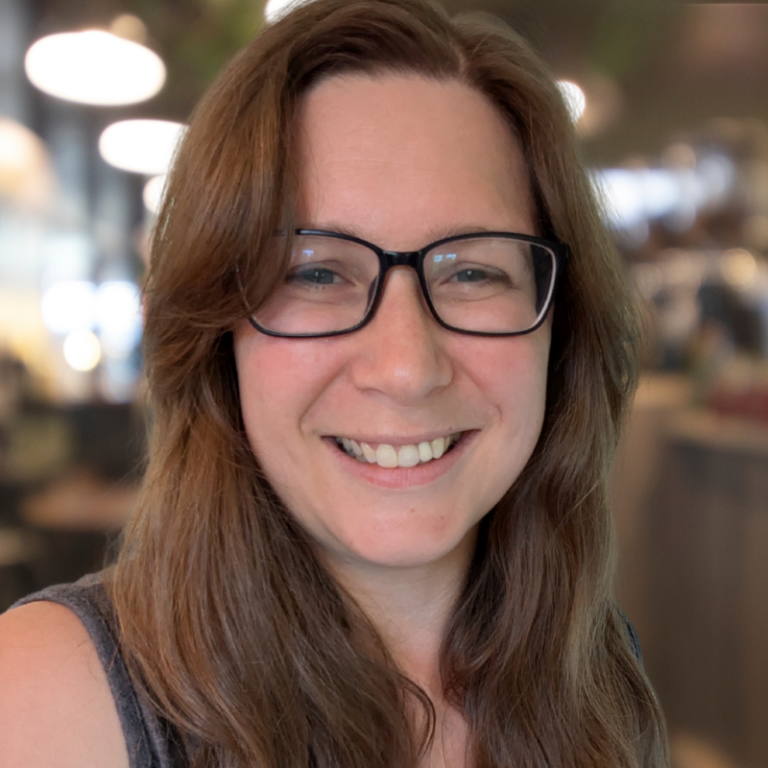 A woman with long brown hair and glasses smiling indoors.