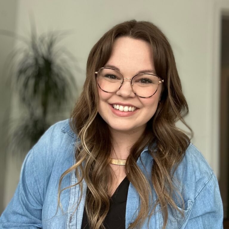 A young woman with long brown wavy hair, glasses, and a bright smile, wearing a denim jacket and gold necklace, standing indoors with a plant in the background.