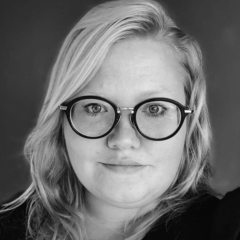 A black and white close-up portrait of a woman with light-colored hair and round glasses, smiling slightly.