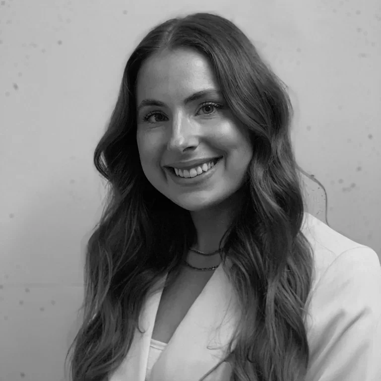 A young woman with long wavy hair smiling, wearing a light-colored blazer and layered necklaces, against a plain background.