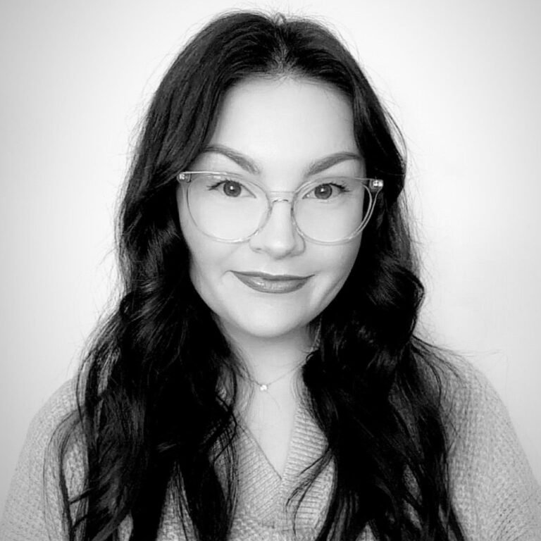 Black and white portrait of a woman with long wavy dark hair, wearing glasses, a V-neck sweater, and a necklace, smiling at the camera.