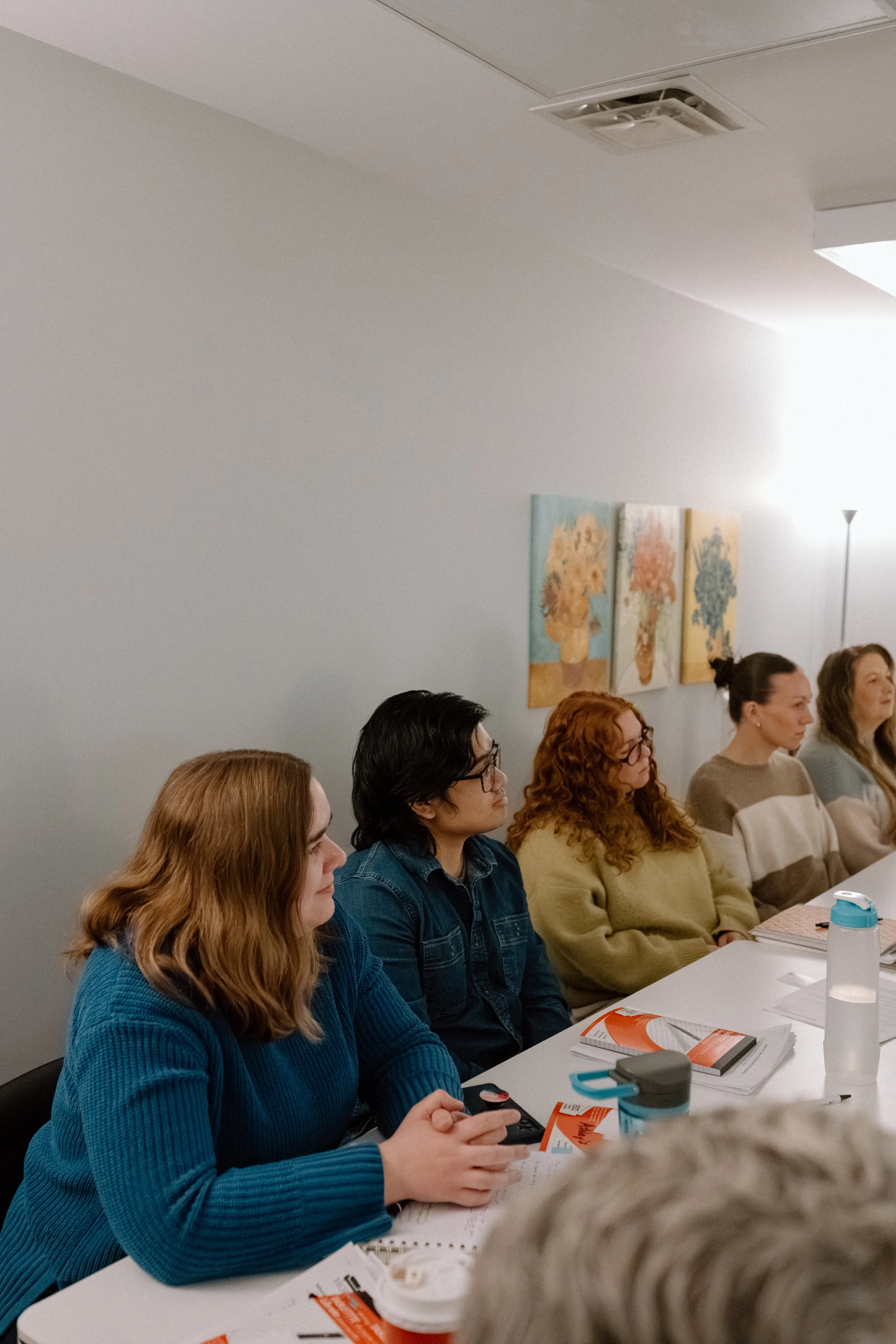 A group of people sitting at a long table in a room with white walls, watching something or listening attentively. The table has notebooks, papers, and drink bottles, and some individuals are wearing glasses, with hair colors including blonde, brown, and red. There are three paintings of floral vases on the wall, and a bright light source is visible in the background.
