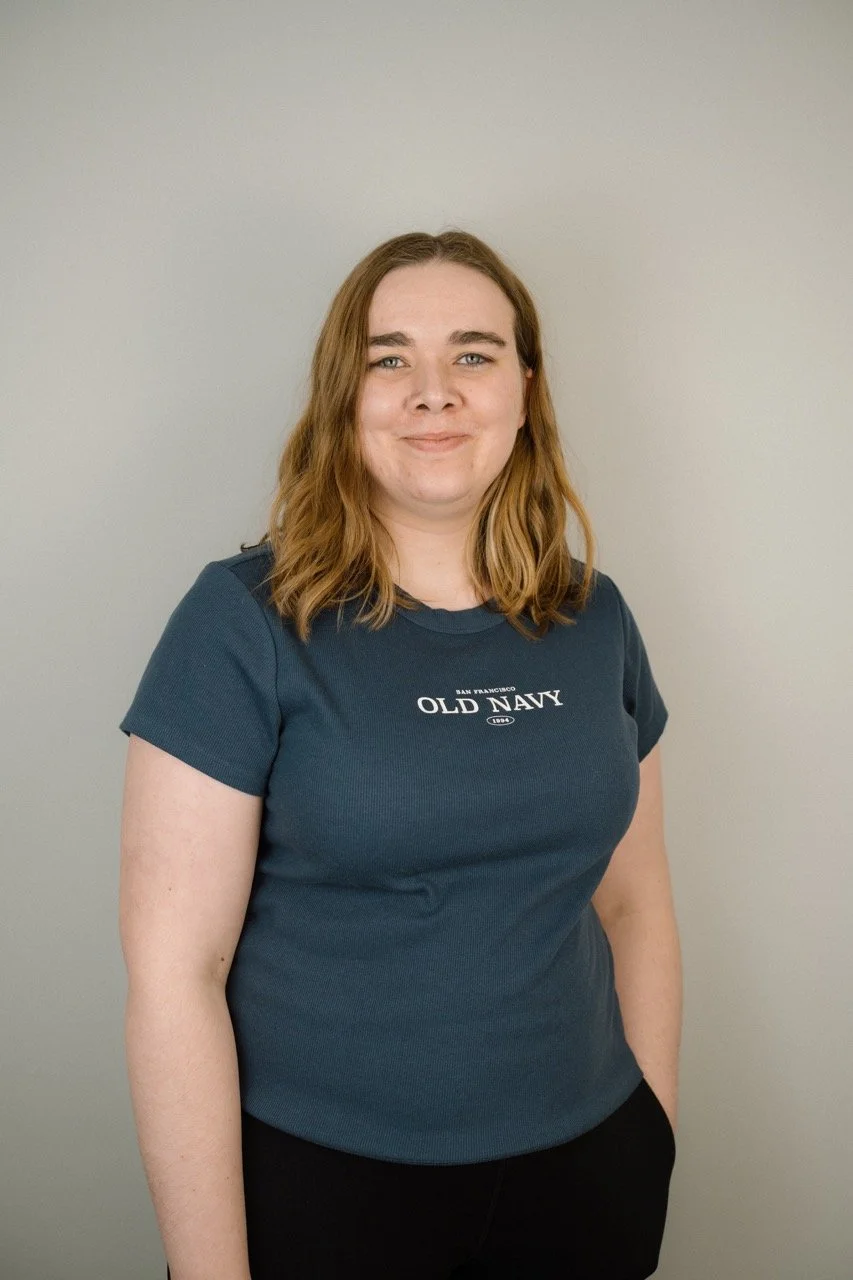 Young woman with shoulder-length wavy brown hair, smiling, wearing a dark blue Old Navy T-shirt and black pants, standing against a plain light gray wall.