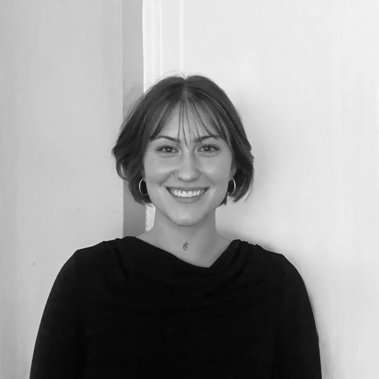 Black and white photo of a smiling young woman with short hair, wearing hoop earrings and a dark top, standing against a plain wall.