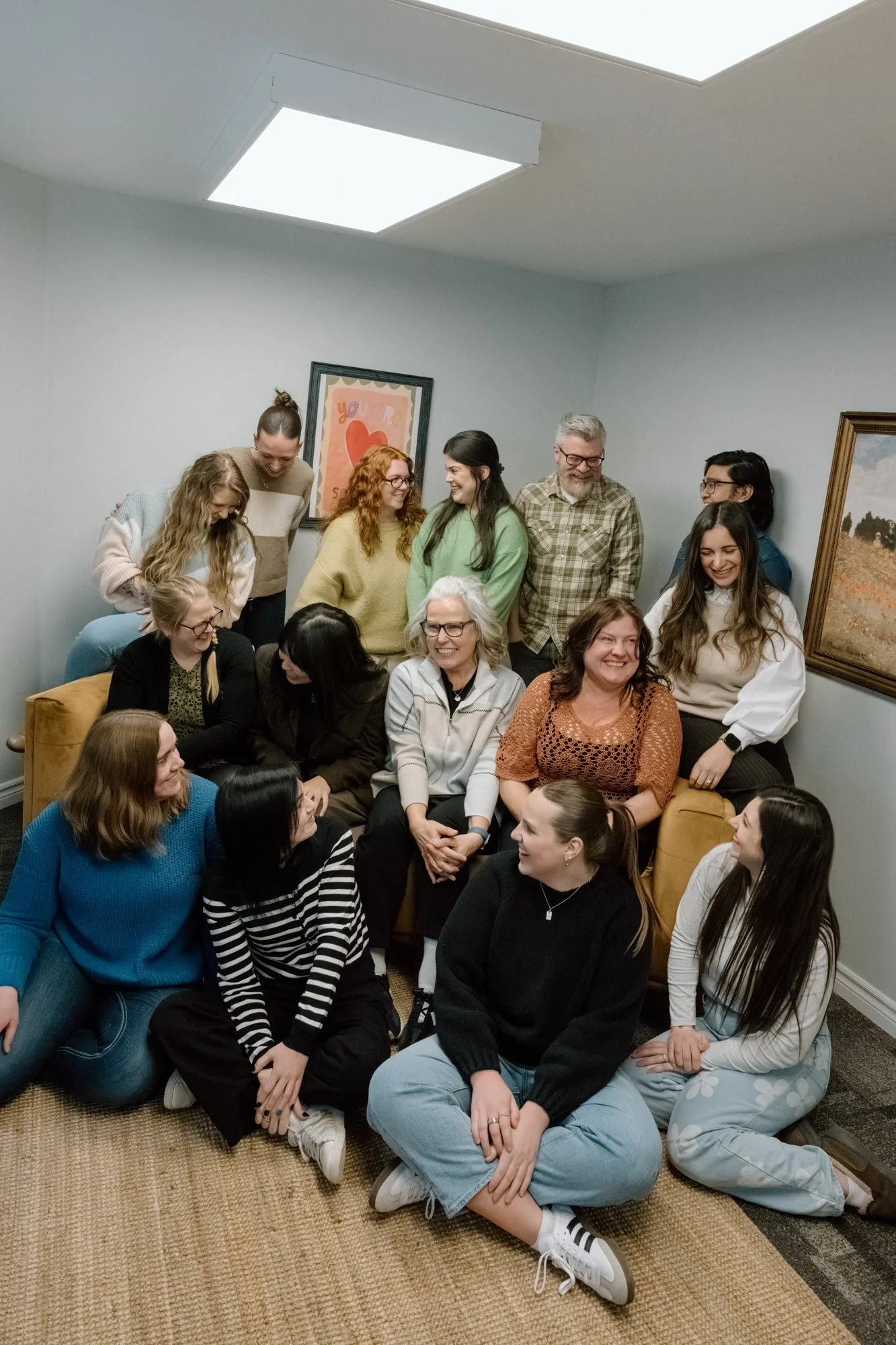 Group of diverse women and men gathering in a cozy, well-lit room, sitting and standing, smiling and engaging in conversation.