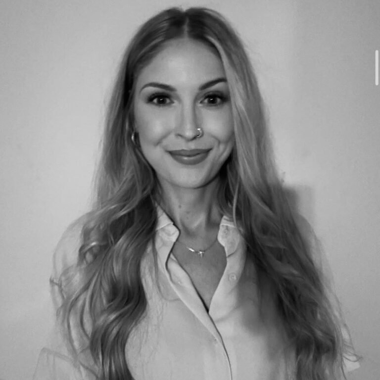 A woman with long wavy hair, wearing a collared shirt and jewelry, smiling at the camera in front of a plain wall.