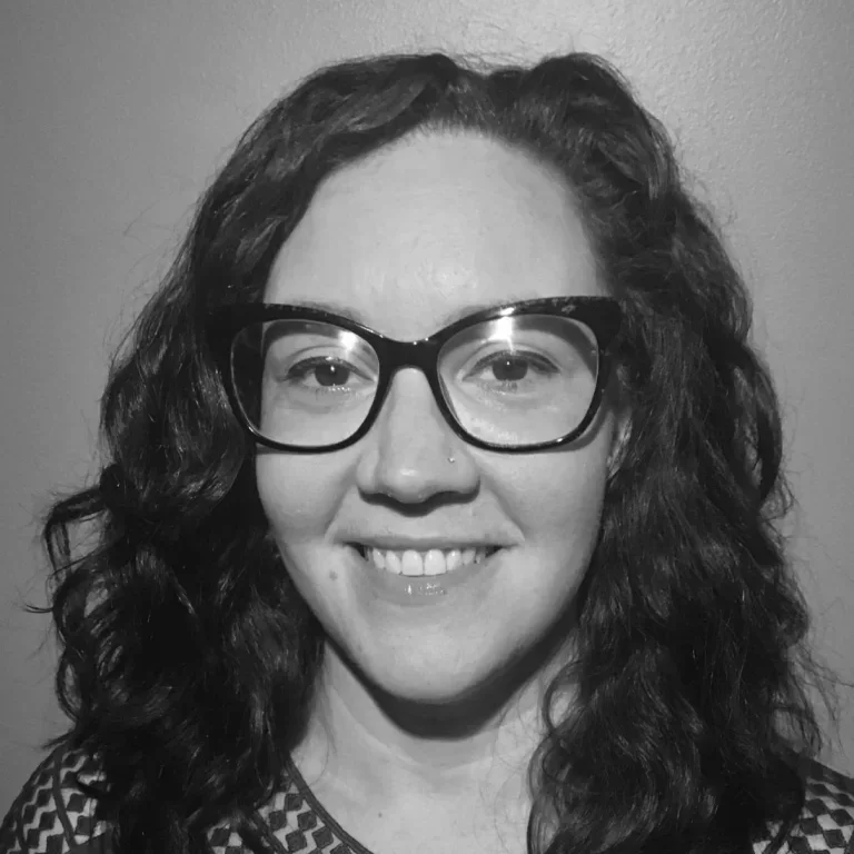 A woman with curly hair and glasses smiling at the camera in black and white.