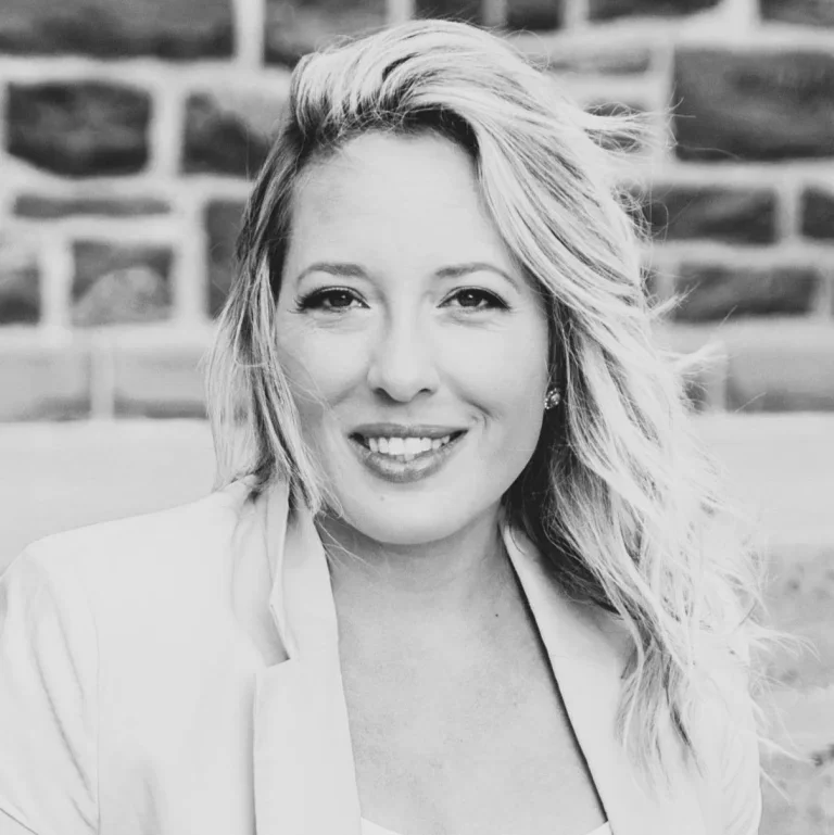 Close-up black and white portrait of a smiling woman with wavy hair, wearing a blazer, outdoors with a brick wall in the background.