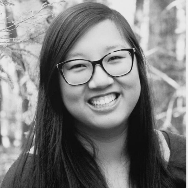A smiling woman with long dark hair and glasses outdoors in a natural setting.