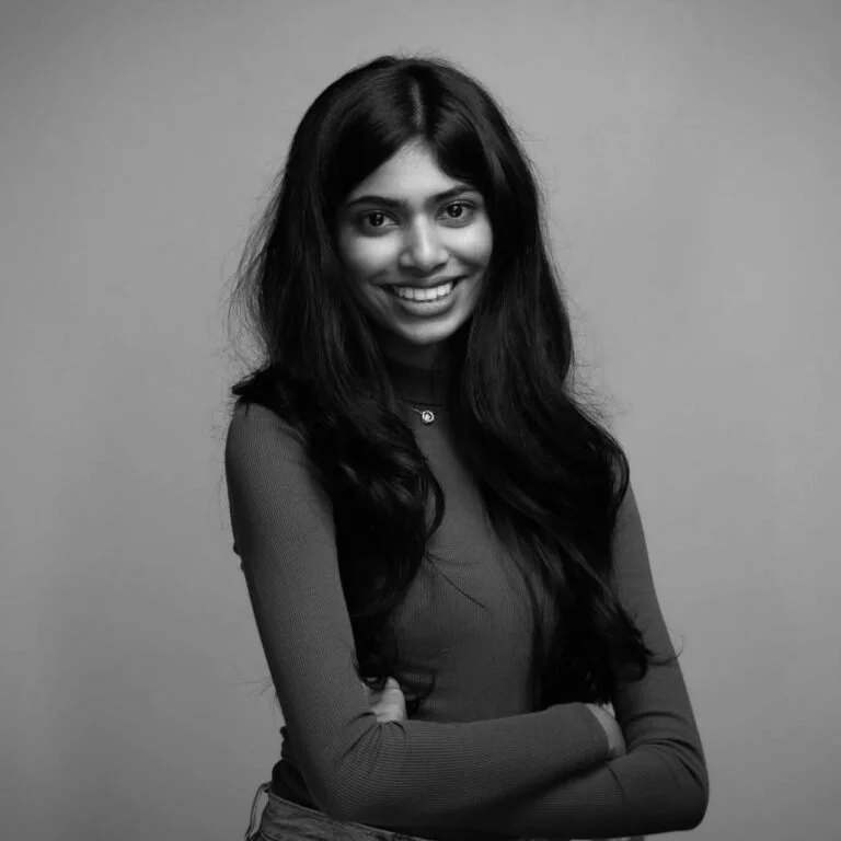 Black and white portrait of a young woman with long wavy hair, smiling, wearing a long-sleeve top, standing in front of a plain background.