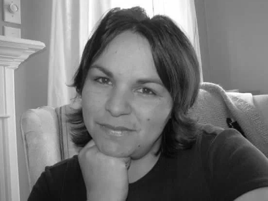 Black and white photo of a woman with medium-length hair sitting on a couch, resting her chin on her hand and smiling slightly.