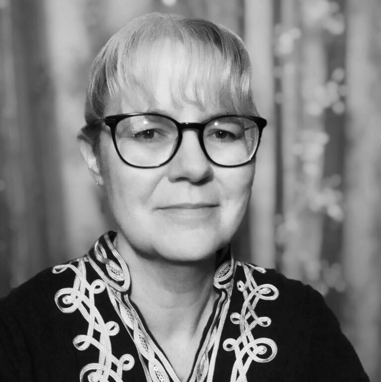 A woman with short blonde hair, glasses, and a patterned shirt, smiling slightly in a black and white photo with a blurred background.