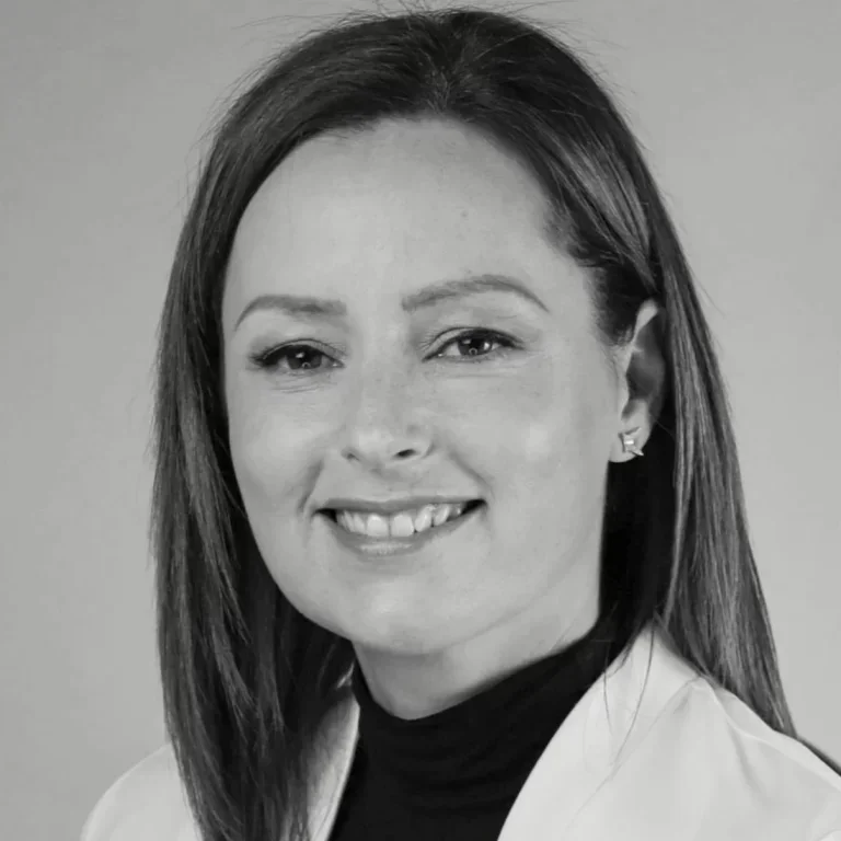 Black and white portrait of a woman with shoulder-length hair, smiling, wearing earrings and a light-colored blazer over a dark turtleneck.