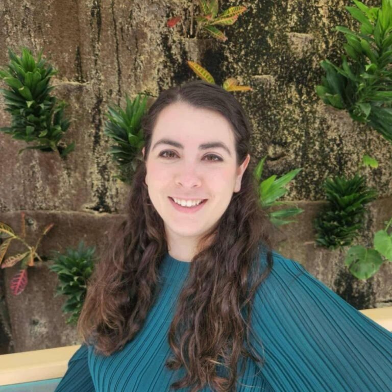 Young woman with long brown curly hair smiling, wearing a teal pleated top, standing in front of a textured stone wall with green plants and small yellow flowers.