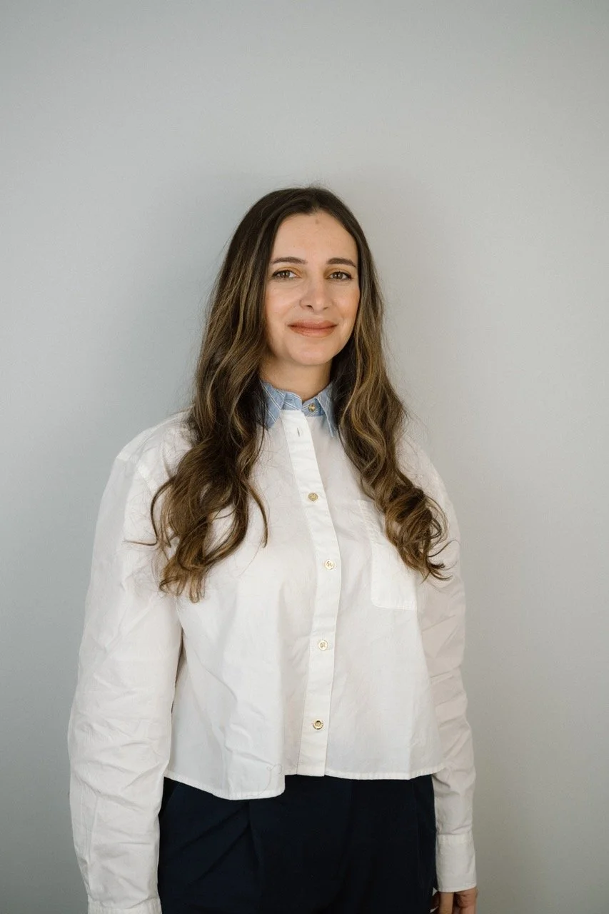 A woman with long, wavy brown hair standing against a plain light gray wall, wearing a white button-down shirt with a light blue collar and dark pants.