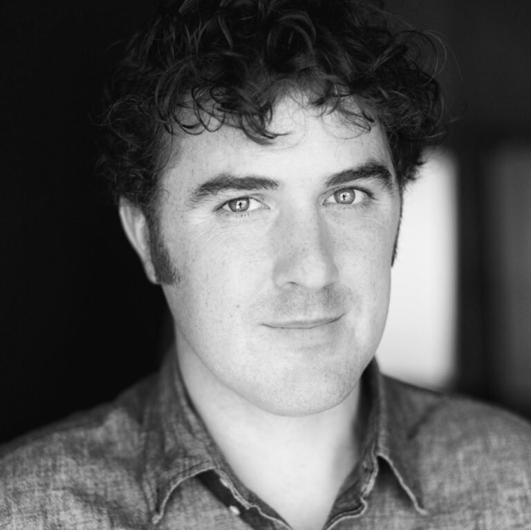 Black and white portrait of a young man with curly hair, light eyes, and wearing a denim shirt, smiling softly at the camera.