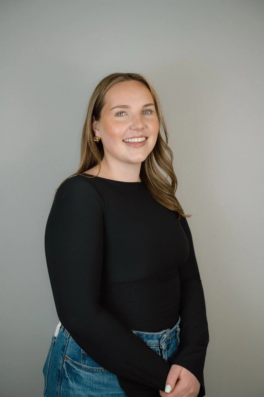 A young woman with light brown hair, wearing a black long-sleeve shirt and blue jeans, standing against a plain gray background and smiling at the camera.