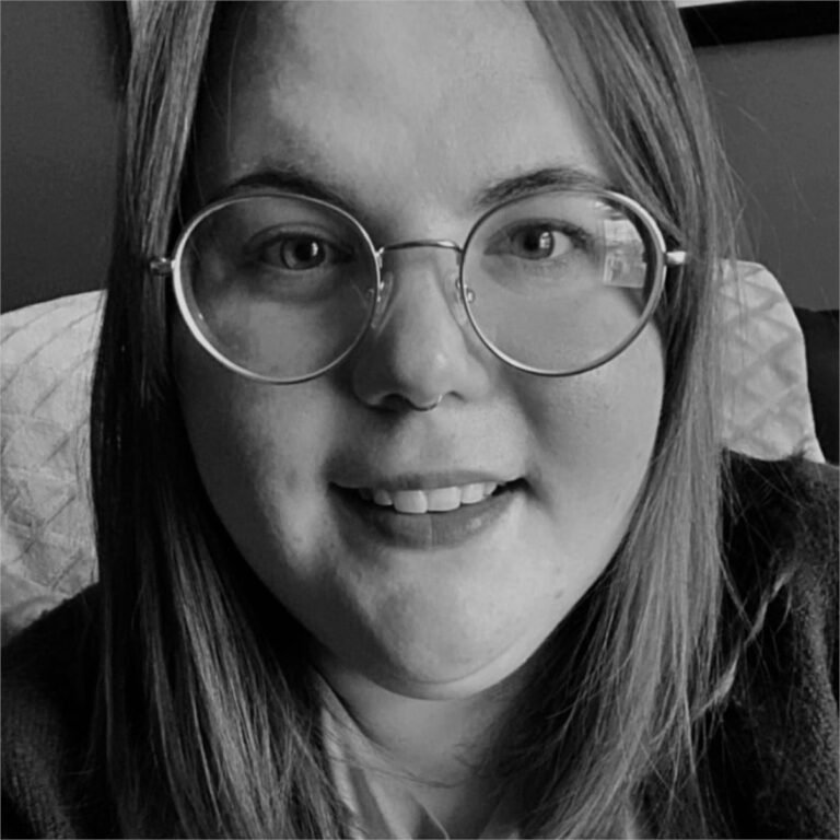 Close-up black and white photo of a person with straight hair, wearing round glasses, smiling with teeth slightly showing, sitting on a bed or couch with a textured pillow in the background.
