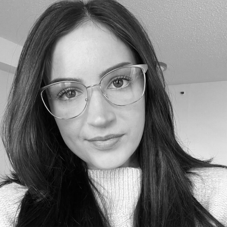 A young woman with long dark hair wearing glasses and a light-colored sweater, looking at the camera with a slight smile, indoors.