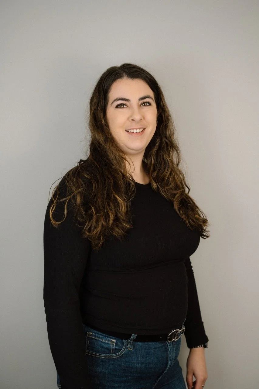 A woman with long, curly brown hair, wearing a black long-sleeve shirt and blue jeans, smiling and standing against a plain gray wall.