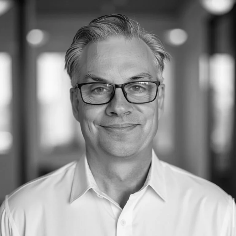 Black and white portrait of a middle-aged man with glasses, smiling, wearing a white collared shirt, in an office or professional setting.