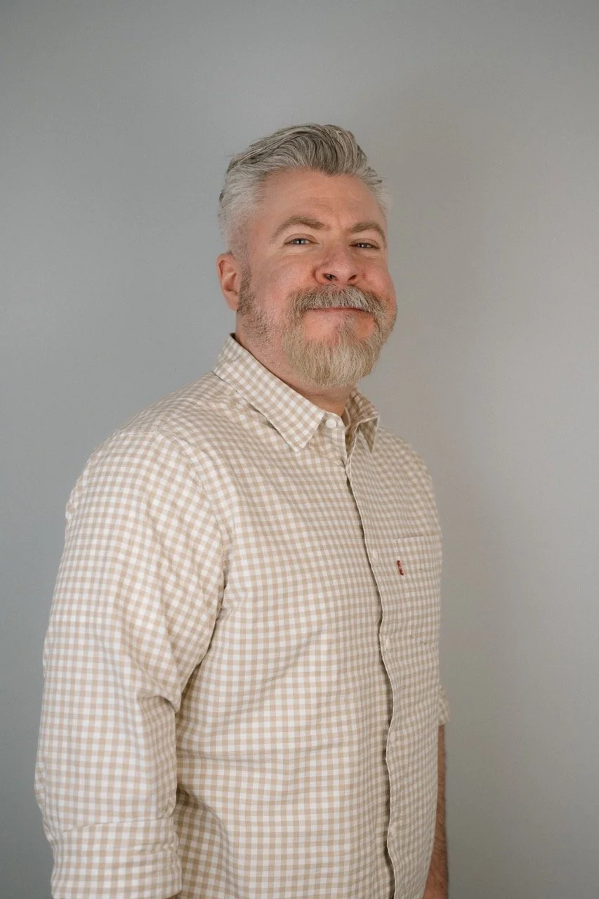 Portrait of a smiling man with gray hair and beard, wearing a beige checkered shirt, standing against a plain gray background.