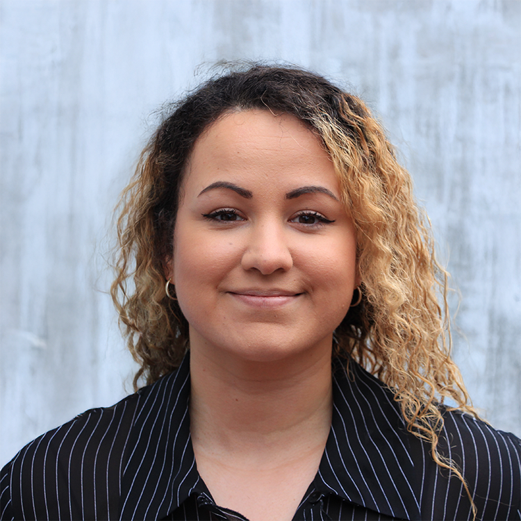A woman with curly blonde hair wearing a black pinstripe blazer, smiling against a gray concrete wall background.