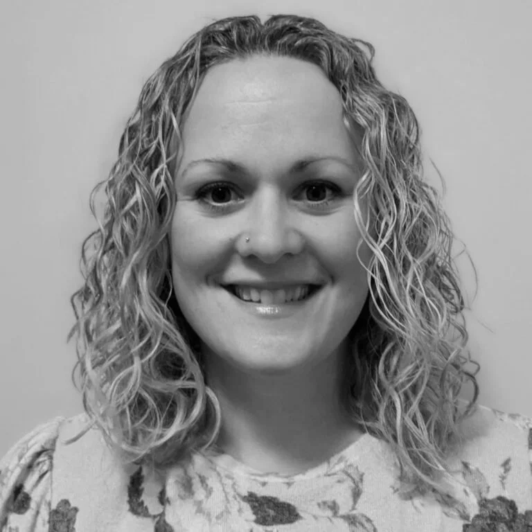 A smiling woman with curly hair and a nose piercing wearing a floral patterned top, standing against a plain background.