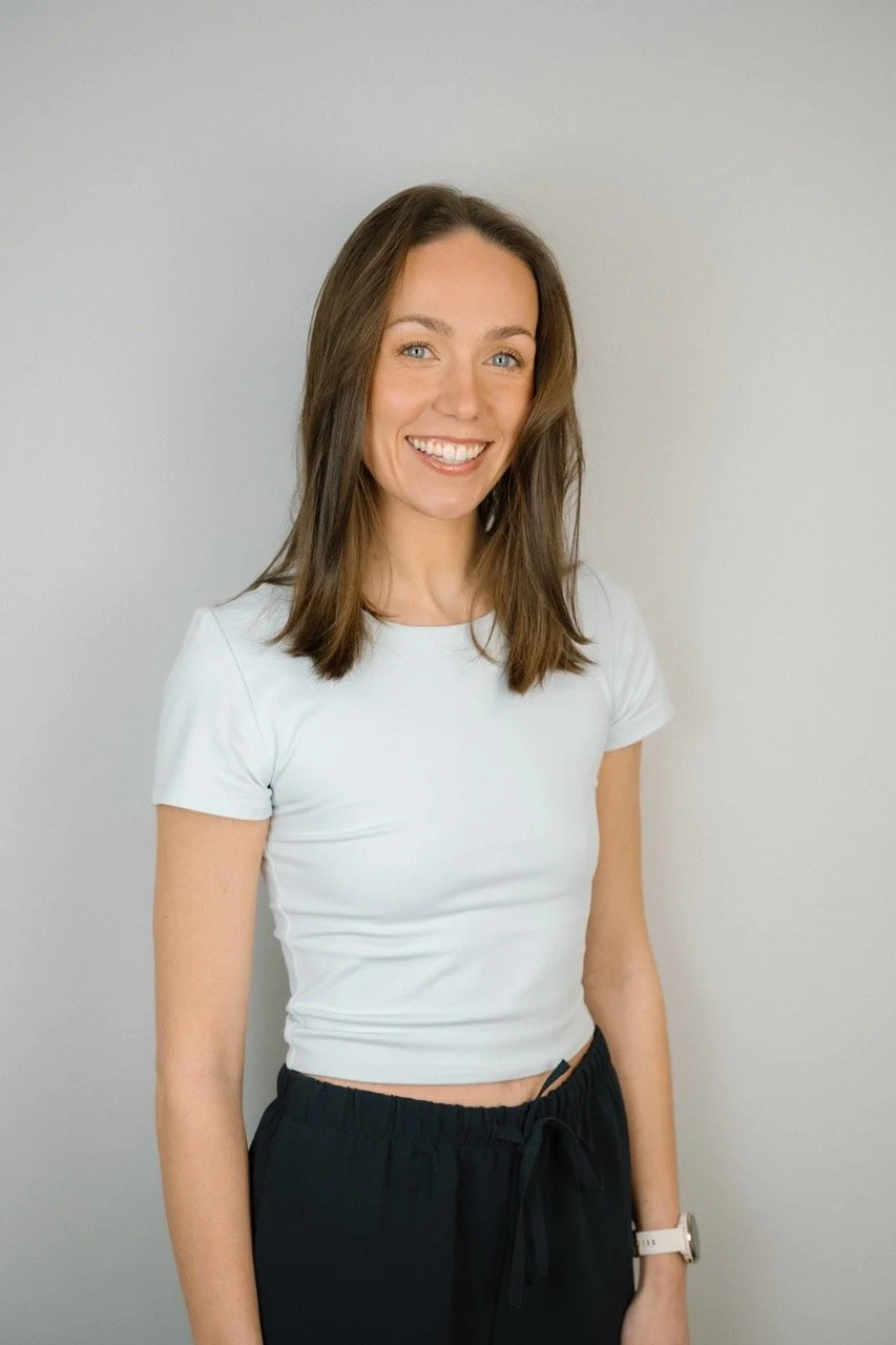 A woman with shoulder-length brown hair, blue eyes, and fair skin smiling, wearing a white t-shirt and black drawstring pants, standing against a light-colored wall.