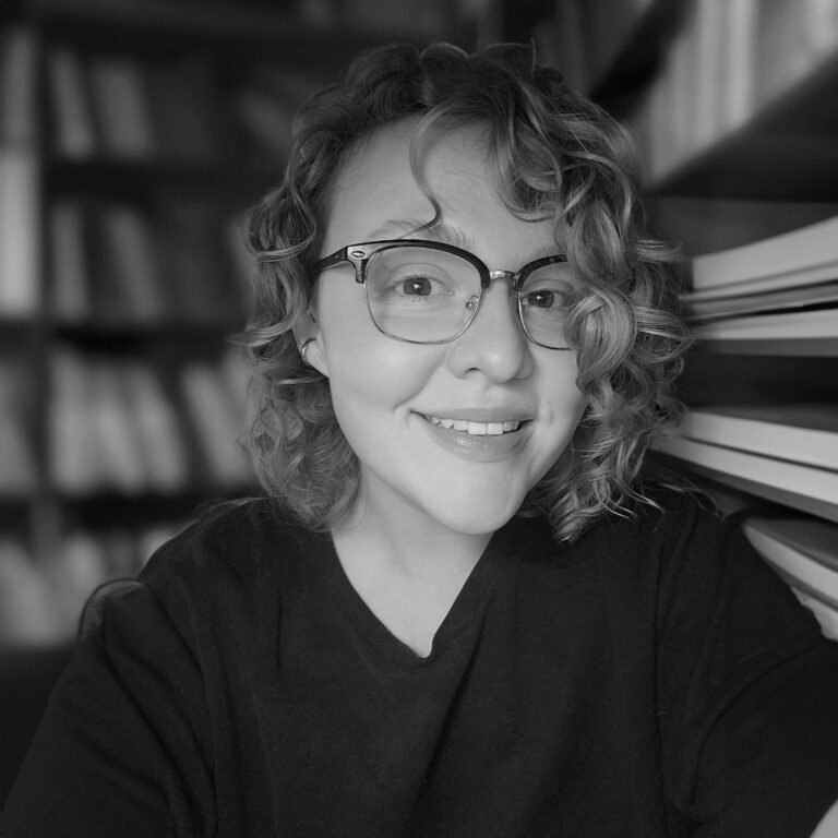 A woman with curly hair and glasses smiling at the camera in front of bookshelves and a stack of books.