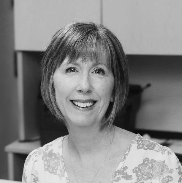 Black and white photo of a smiling woman with short hair and a patterned blouse, indoors with cabinets in the background.