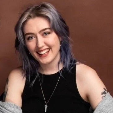 Smiling young woman with shoulder-length wavy hair dyed purple and silver, wearing a black sleeveless top and a necklace, sitting against a brown background.