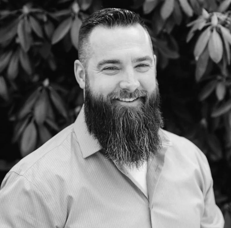 A smiling man with a full beard and short hair, wearing a collared shirt, standing outdoors in front of leafy bushes.