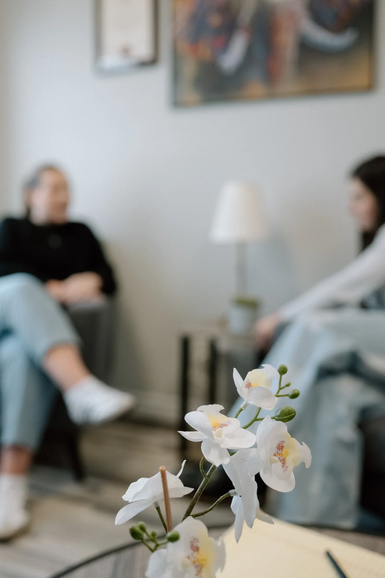 Close-up of white orchids with pink and yellow centers on a vase in the foreground, with blurred people sitting and chatting in a room in the background.