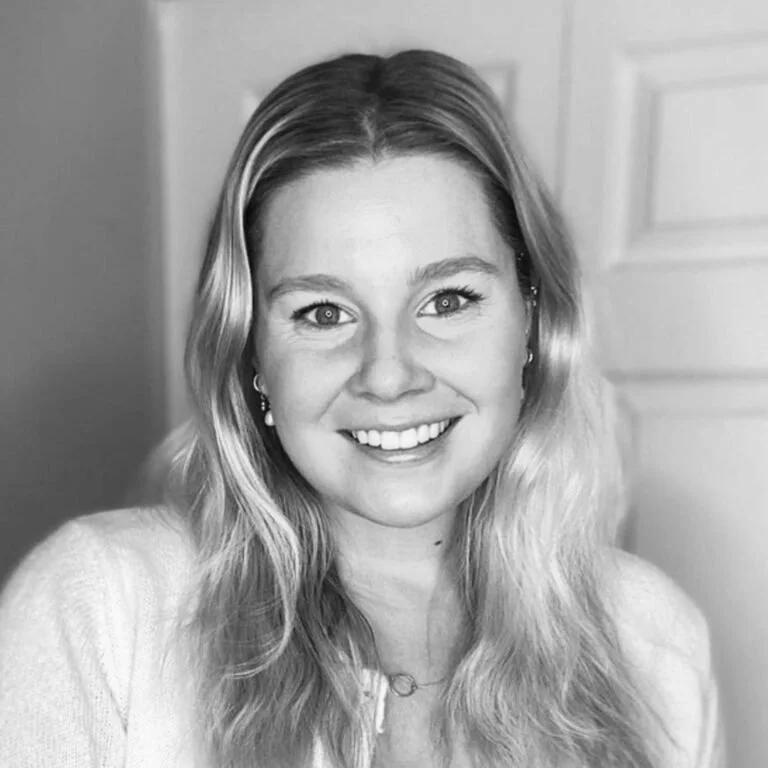 A smiling woman with wavy blonde hair, wearing earrings, a light-colored top, and a necklace, in front of a plain background.