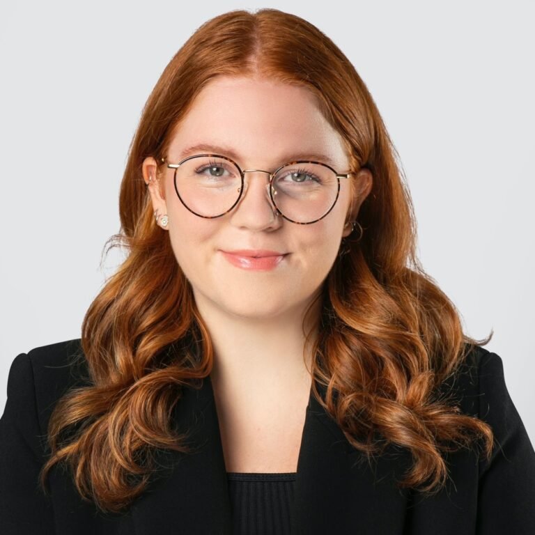 A woman with long, wavy red hair, wearing round glasses and a black blazer. She has a slight smile and is looking directly at the camera.